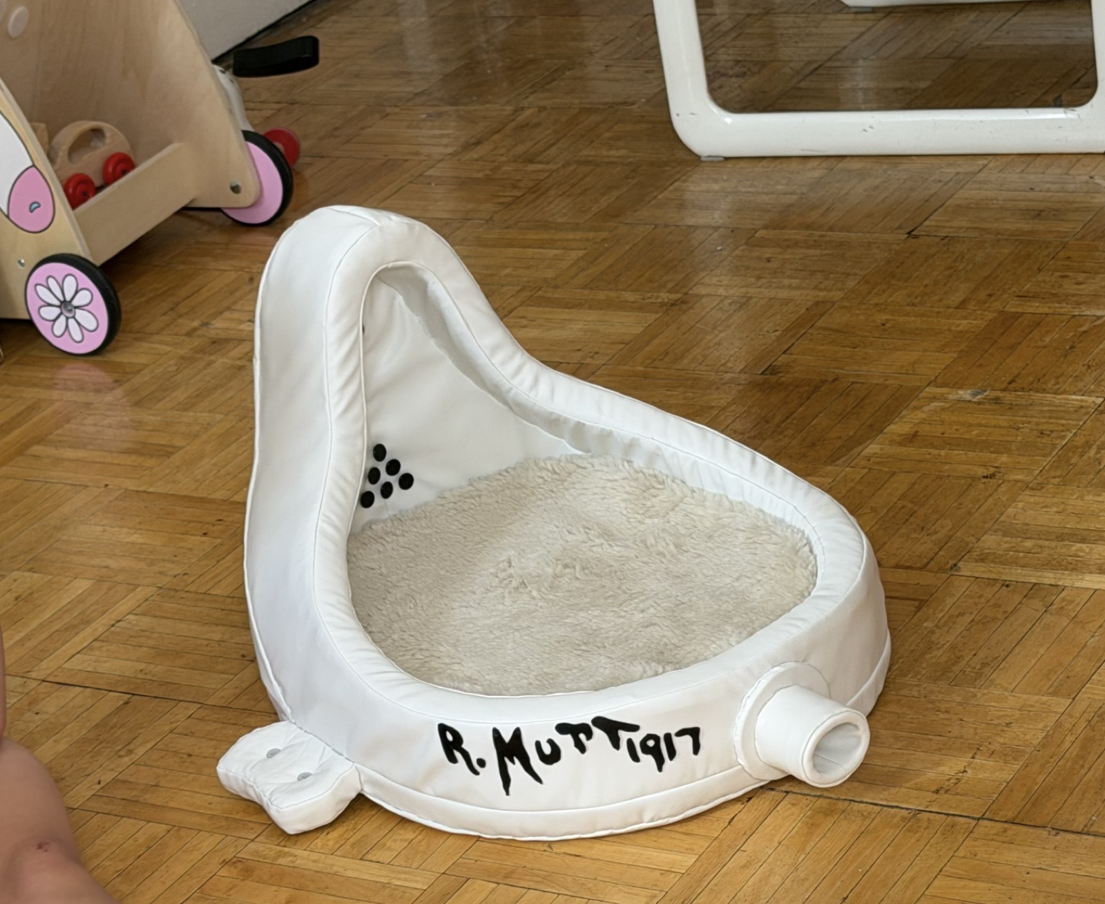 The plush white dog bed in the shape of Duchamp's urinal, signed R. Mutt 1917, on a parquet floor at home